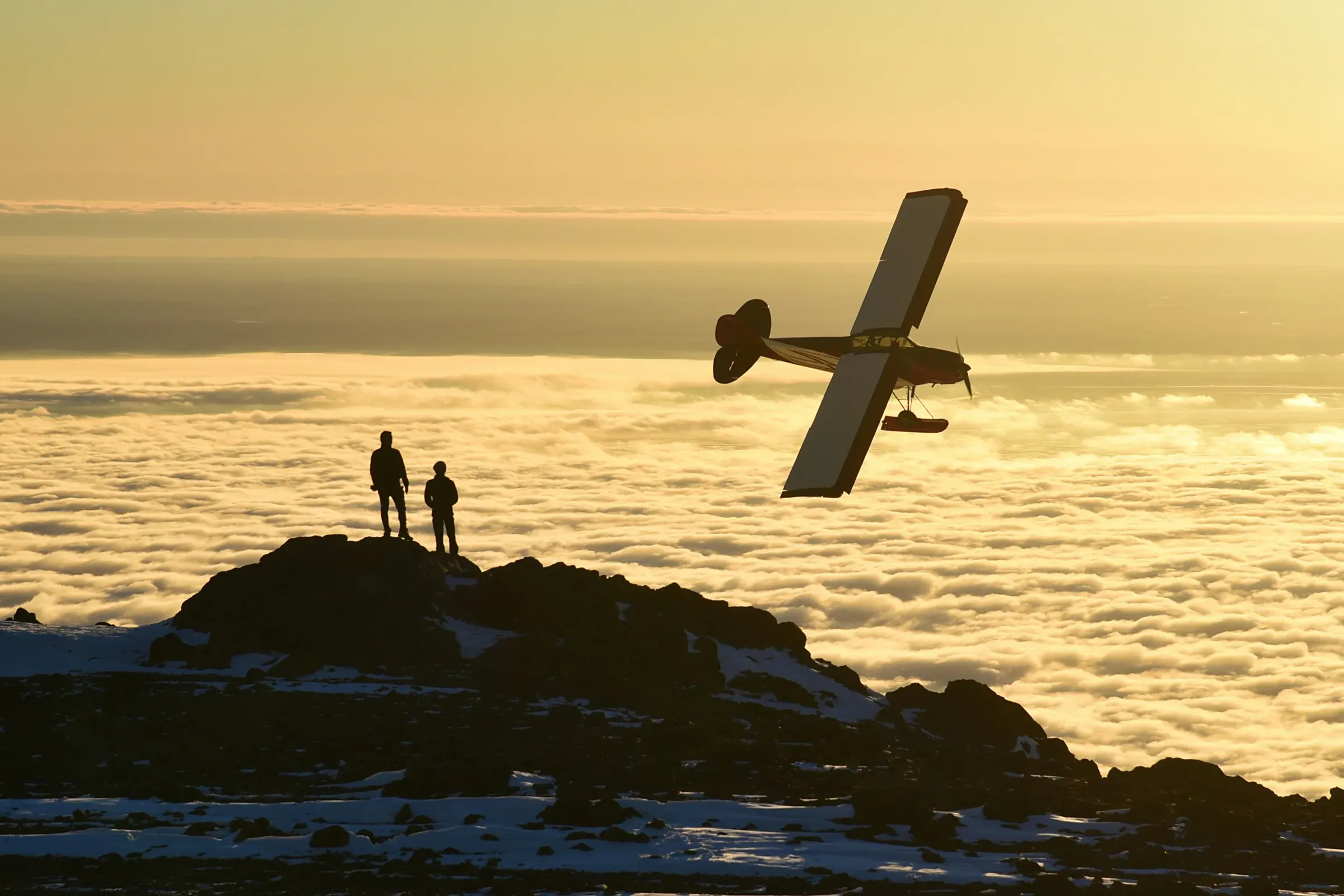 A small plane flying over a sea of clouds at sunset, watched by two people on a rocky summit